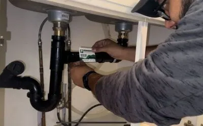 A person wearing glasses and a cap works under a sink, using a card to inspect the black plumbing pipes and connections—a meticulous approach often seen in Queen Creek plumbing services.