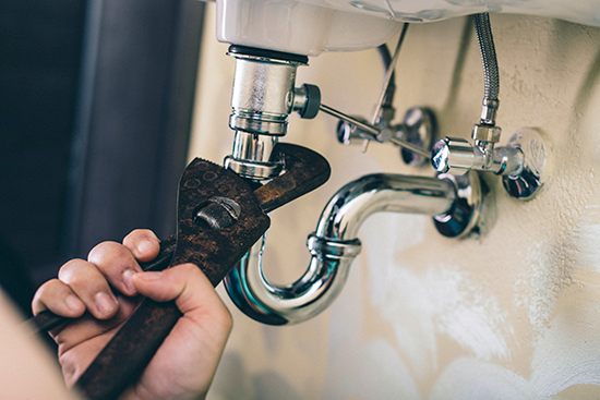A person uses a wrench to tighten or loosen a pipe under a bathroom sink, handling plumbing repairs with shiny metal pipes and connectors—typical of professional Apache Junction Plumbing services.
