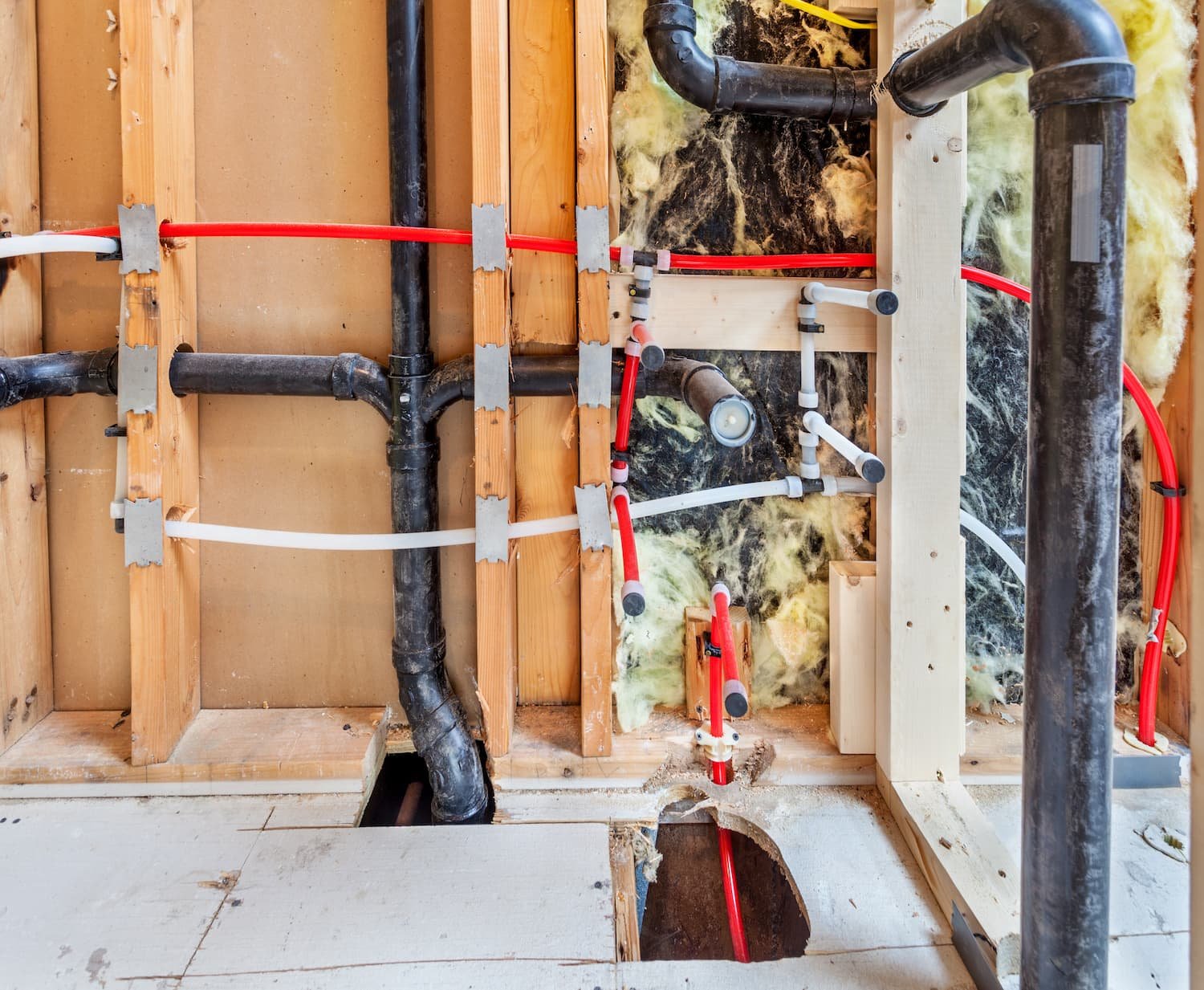 Exposed wall with plumbing pipes, black drain pipes, red and white PEX water lines, yellow insulation, and wood framing. A hole is cut in the floor for pipe access—perfect for Apache Junction Plumbing repairs or upgrades.